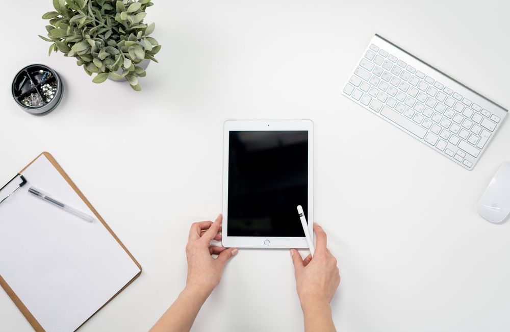 Persona che scrive con la penna su un tablet appoggiato a una scrivania, con tastiera e taccuino